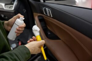 Person spraying cleaning foam onto a brush, preparing to clean the interior door panel of a car with brown and black trim—a meticulous step in professional Car Detailing Services in Kenner.