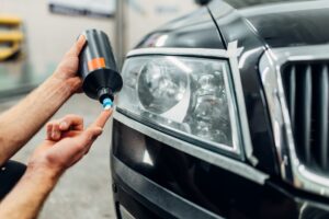A mechanic in uniform inspects the headlight of a white car with the hood open, checking for issues or performing Headlight Restoration New Orleans-style maintenance outdoors.