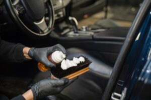A person wearing black gloves applies cleaning foam to a brush inside a car, preparing to clean the vehicle's interior—showcasing the precision of Car Detailing Services in Kenner.