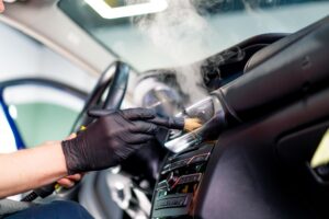 Wearing black gloves, a person uses a brush and steam cleaner to clean the dashboard and air vents inside a car—showcasing the quality you can expect from top mobile detailing services in LaPlace.