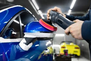 A person performs Auto Paint Correction in New Orleans, using a power buffer to polish the side mirror of a blue car, with masking tape applied for protection, inside a well-lit garage.