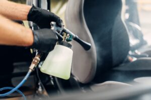 A person wearing black gloves uses a spray cleaning tool for meticulous auto detailing in Slidell, focusing on the car seat and surrounding interior area.