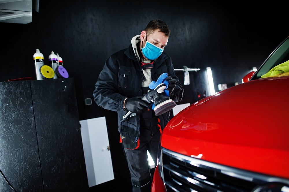 A man wearing a mask and gloves uses a polishing tool to buff the hood of a red car in a Mobile Auto Detailing in Mandeville workshop.
