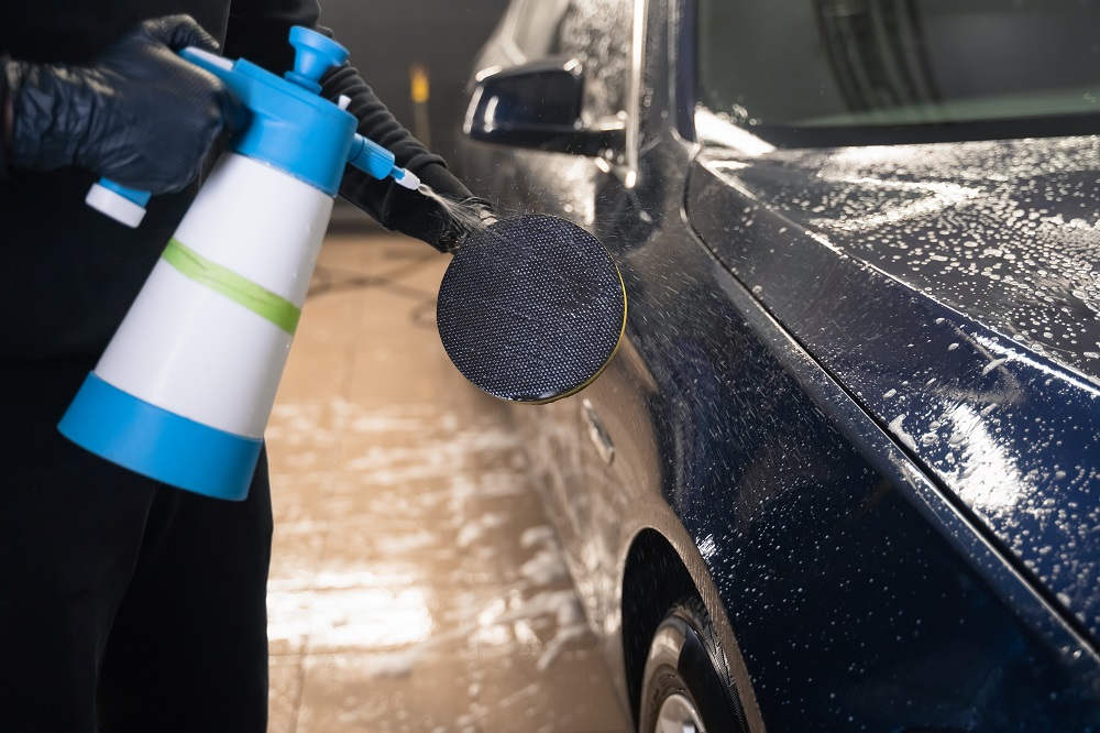 Wearing gloves, a person uses a spray bottle and foam pad to clean the soap-covered exterior of a car—showcasing expert Mobile Exterior Detailing in New Orleans.