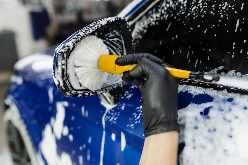 A gloved hand uses a brush to clean the soapy side mirror of a blue car during a Mobile Exterior Detailing in New Orleans.