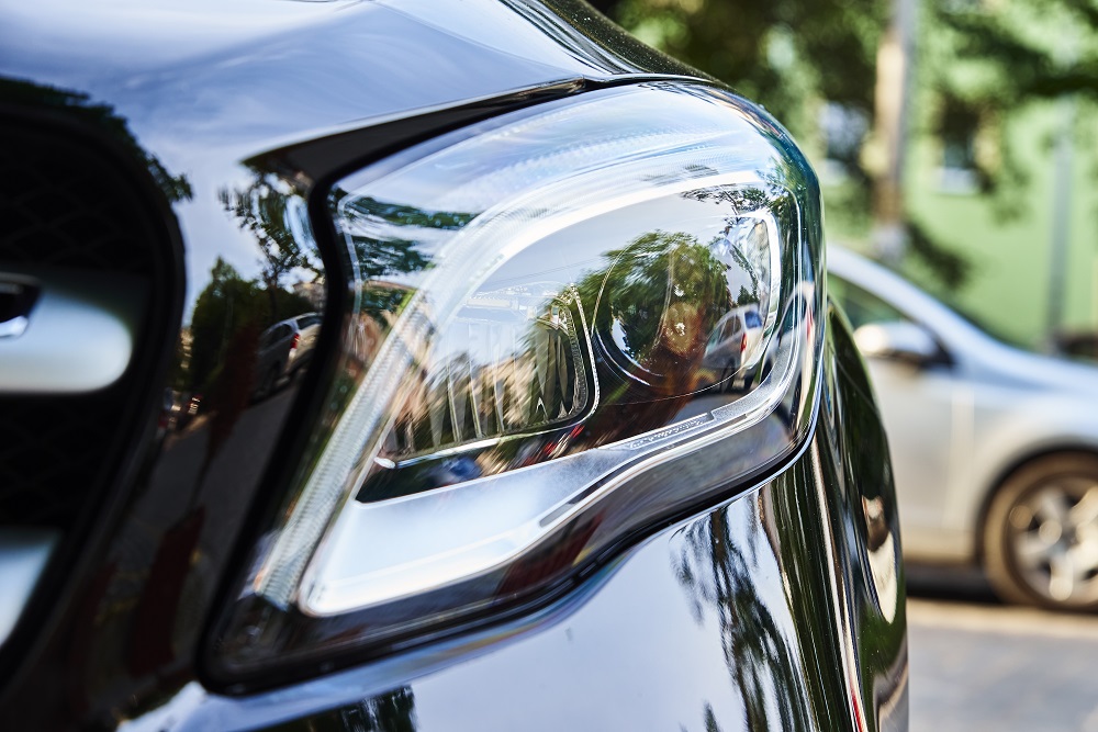 Close-up of the front headlight on a black car in Metairie, with reflections of trees and another parked vehicle—showcasing the shine you get from expert Mobile Auto Detailing in Metairie.