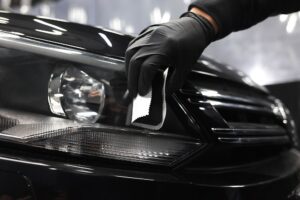 A person wearing a black glove cleans the headlight of a black car with a white cloth, showcasing Headlight Restoration New Orleans.