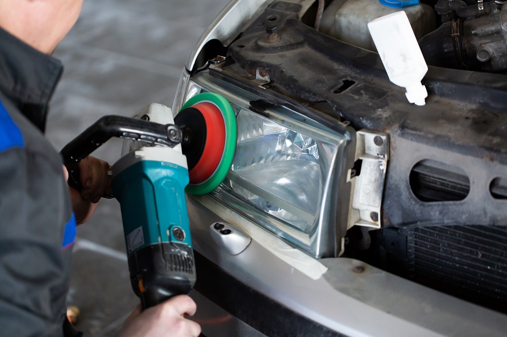 A person uses a power buffer to polish the lens of a car headlight, with the hood open and engine components visible during a Headlight Restoration New Orleans service.