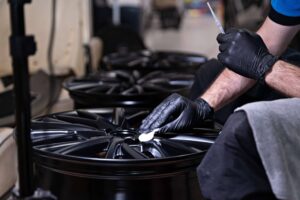 A person wearing black gloves cleans a black alloy wheel with a cloth, preparing it for ceramic coating; several similar wheels are visible in the background.