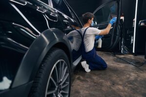 A man wearing gloves and a mask is cleaning the interior of a car door with a vacuum in an Auto Detailing Covington garage or workshop setting.