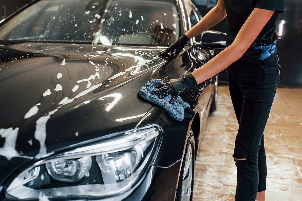 A professional wearing black gloves washes a black car with a soapy cloth, focusing on the hood and headlight area indoors—showcasing expert Auto Detailing in Madisonville.