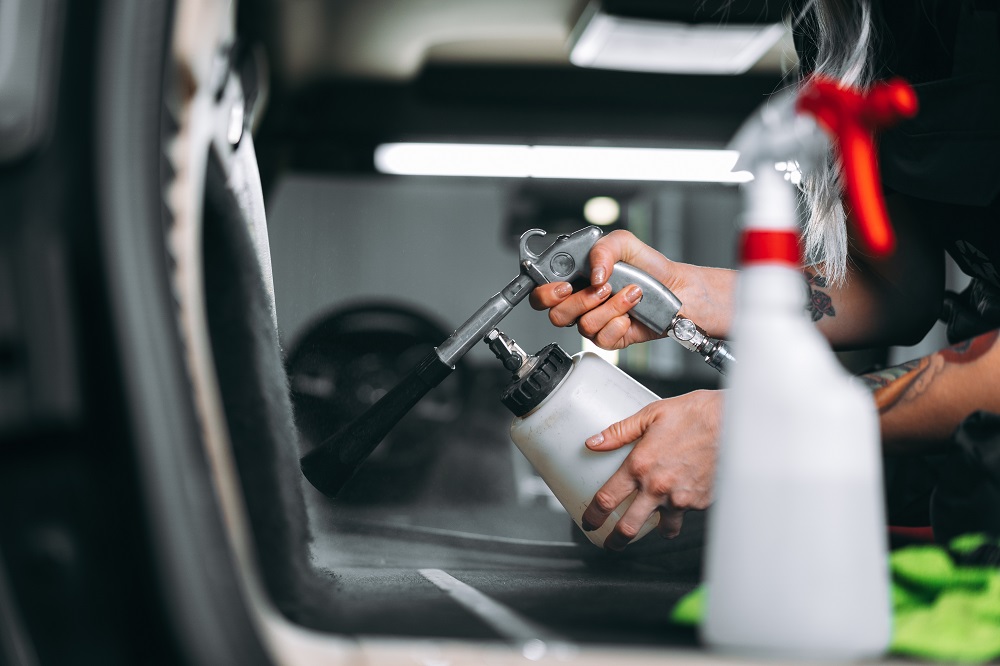 A person performs Auto Detailing in Madisonville, using a spray cleaning tool on a car interior, with a white bottle and green cloth nearby.