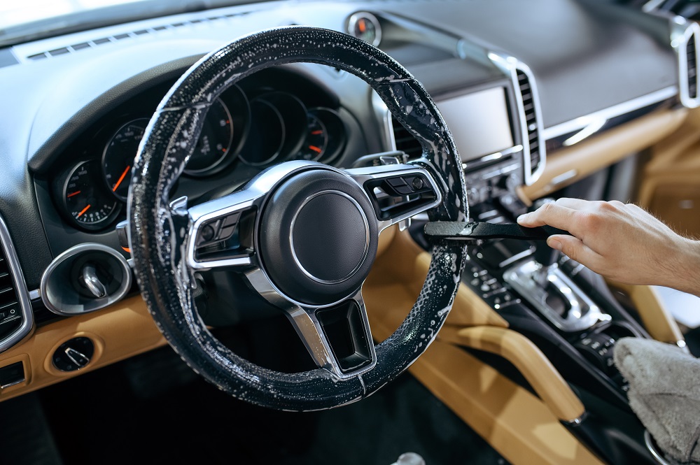 A person cleaning a car’s steering wheel with a brush and cleaning solution showcases expert Auto Detailing in Madisonville; the modern interior features stylish tan and black elements.
