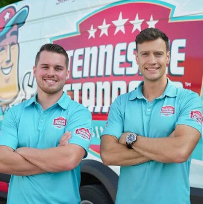 Bobby and Kelton wearing light blue polo shirts stand with arms crossed in front of a Tennessee Standard-branded van.